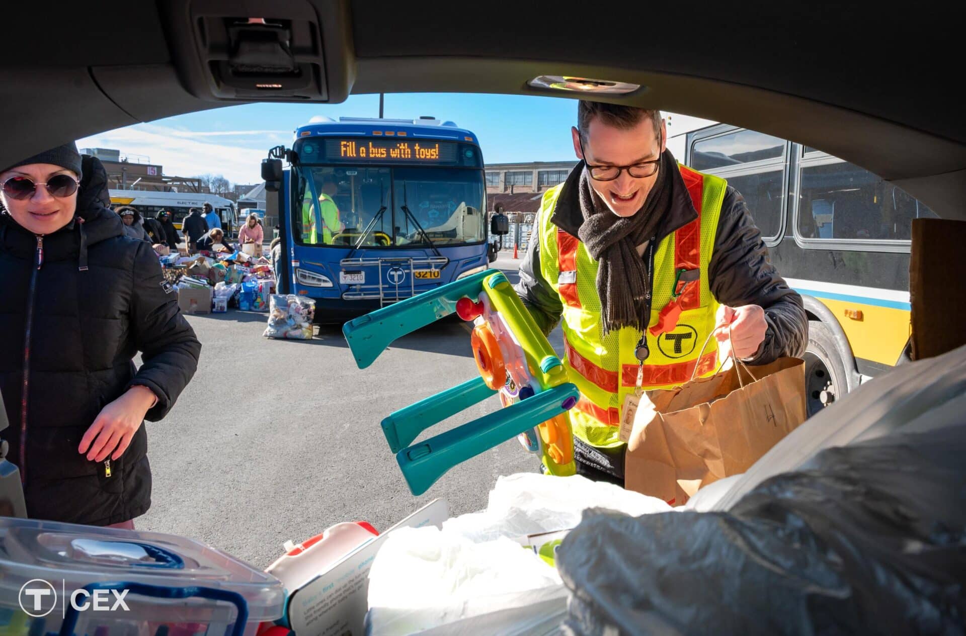 MBTA staff loading toys into a car for transport. A bus with a sign reading "Fill a bus with toys!" is in the background.